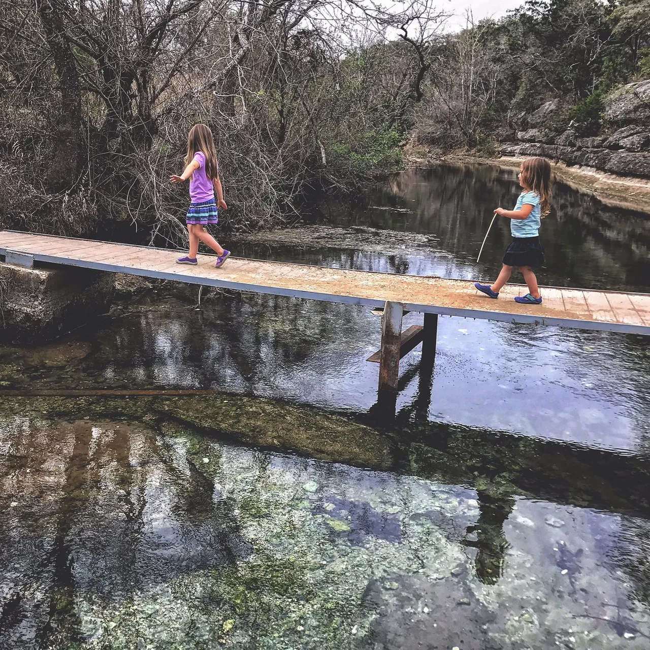 Two children cross a narrow footbridge over still water.