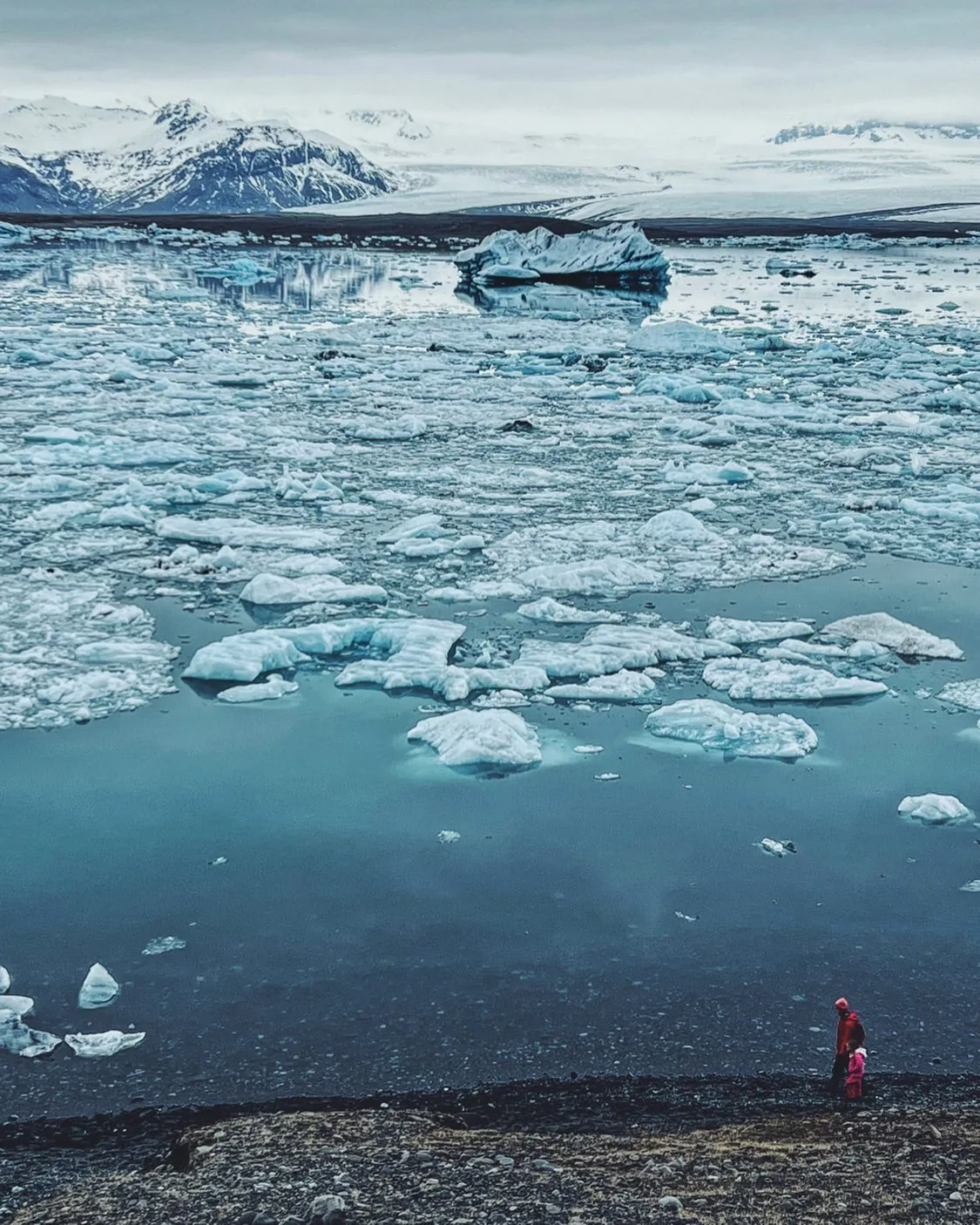 Blue ice drifts across a lagoon beneath snow-covered mountains.