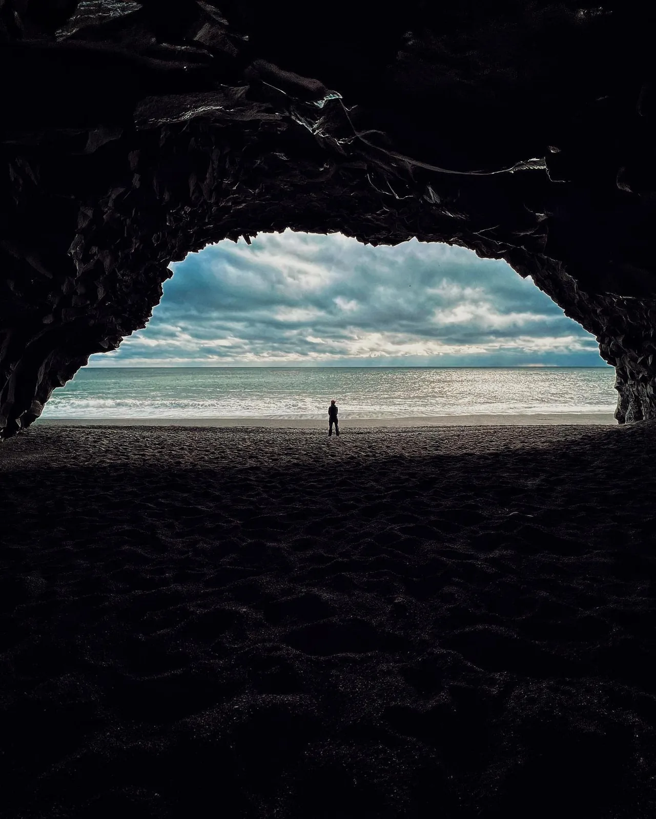 A person stands on a beach framed by the dark mouth of a sea cave in Iceland.